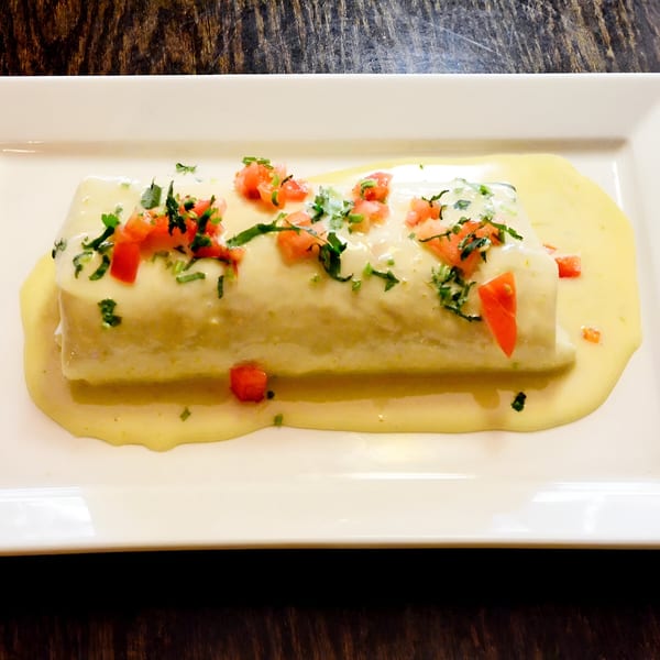 A Mexican burrito covered in creamy yellow sauce, topped with diced tomatoes and fresh cilantro.